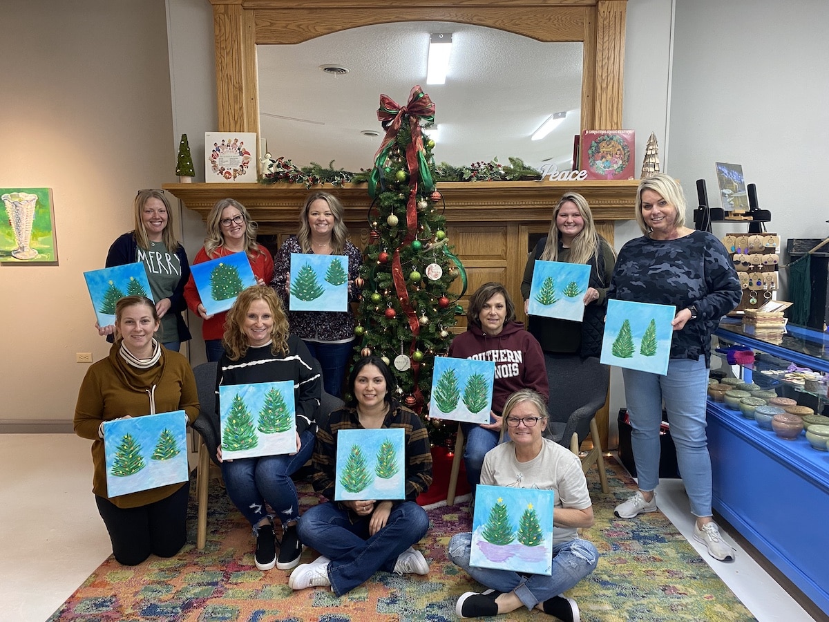 Women holding paintings of pine trees in front of a Christmas tree